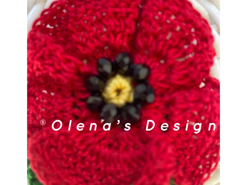May include: Close-up of a handmade red crocheted flower with a yellow center and black bead accents. The flower is part of a larger design, with the text "Olena's Design" visible.