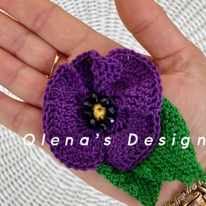 May include: A handmade crochet flower in shades of purple, green, and black. The flower has a detailed design with a beaded center and a green leaf. The text "Olena's Design" is visible.