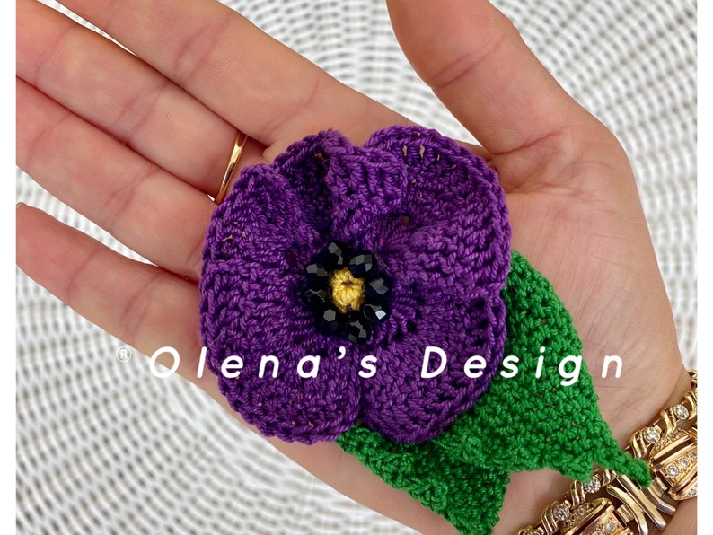 May include: A handmade crochet flower in shades of purple, green, and black. The flower has a detailed design with a beaded center and a green leaf. The text "Olena's Design" is visible.