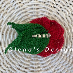 May include: A handmade crochet flower pin with a red flower and green leaf design. The pin has a silver-colored clasp on the back. The text "Olena's Design" is visible on the image. The pin is placed on a white woven surface.