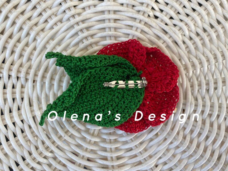 May include: A handmade crochet flower pin with a red flower and green leaf design. The pin has a silver-colored clasp on the back. The text "Olena's Design" is visible on the image. The pin is placed on a white woven surface.