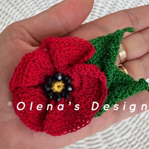 May include: A handmade crochet poppy flower with a green leaf. The red flower has a black and yellow beaded center. The text "Olena's Design" is visible. This floral accessory is perfect for adding a touch of handmade charm.