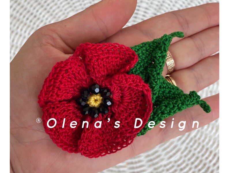 May include: A handmade crochet poppy flower with a green leaf. The red flower has a black and yellow beaded center. The text "Olena's Design" is visible. This floral accessory is perfect for adding a touch of handmade charm.