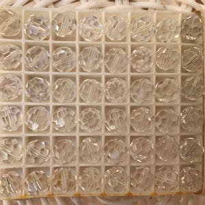 May include: A box of clear, faceted crystal beads. Each bead is round with multiple facets, creating a sparkling effect. The beads are arranged in a grid within a white plastic container. The box has a label with text.