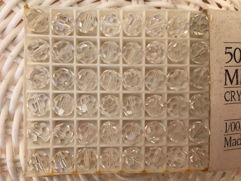 May include: A box of clear, faceted crystal beads. Each bead is round with multiple facets, creating a sparkling effect. The beads are arranged in a grid within a white plastic container. The box has a label with text.