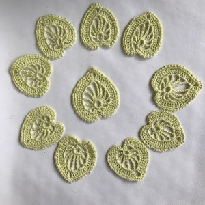 May include: Eleven crocheted leaf-shaped appliques in a pale yellow-green hue. Each applique has an openwork design with a central vein pattern. They are arranged in a circular pattern on a white surface.