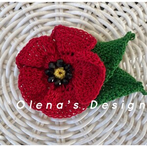 May include: A handmade red crochet poppy flower with a black and yellow center and two green leaves. The flower is set against a white woven background. The text "Olena's Design" is visible.