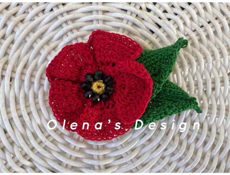 May include: A handmade red crochet poppy flower with a black and yellow center and two green leaves. The flower is set against a white woven background. The text "Olena's Design" is visible.