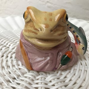 May include: A ceramic frog ornament with a fish-shaped handle. The frog is painted in tan and brown tones, wearing a pink outfit. The fish handle is green, blue, and yellow. An orange and green fishing lure is attached to the frog's outfit.