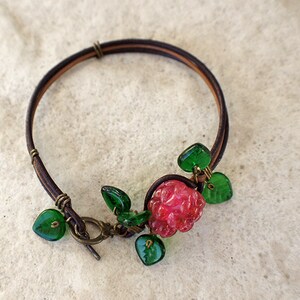 Op de afbeelding: Een bruin leren armband met een rode glazen bloemenbedel en groene glazen bladeren. De armband heeft een eenvoudige sluiting.