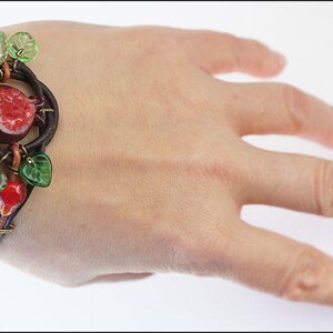 Op de afbeelding: Een bruin leren armband met een rood en groen glazen granaatappelontwerp. De armband zit om een pols.
