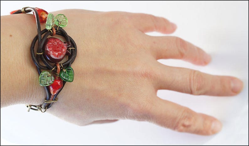 Op de afbeelding: Een bruin leren armband met een rood en groen glazen granaatappelontwerp. De armband zit om een pols.