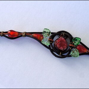Op de afbeelding: Een bruin leren armband met een rood en groen glazen kralenontwerp. De armband heeft een schuifgesp.