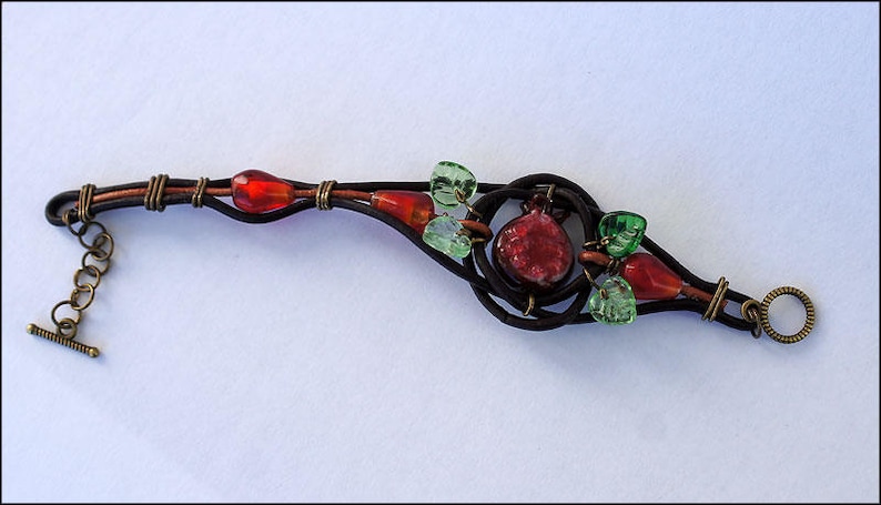 Op de afbeelding: Een bruin leren armband met een rood en groen glazen kralenontwerp. De armband heeft een schuifgesp.