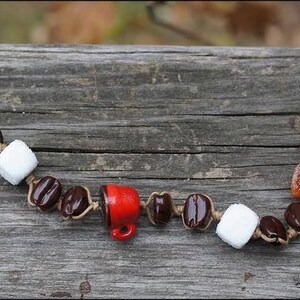 May include: A brown leather bracelet with brown, white, and red beads. The beads are shaped like coffee beans, marshmallows, and a coffee cup.