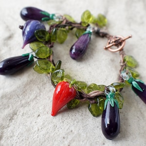 Op de afbeelding: Een armband gemaakt van bruin koord met groene glazen kralen en paarse en rode glazen bedels in de vorm van aubergines en pepers.