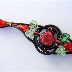 Op de afbeelding: Een bruin leren armband met rode en groene glazen kralen, waaronder een rode glazen granaatappelbedel.