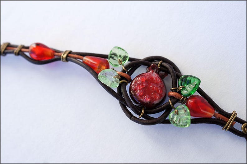 Op de afbeelding: Een bruin leren armband met rode en groene glazen kralen, waaronder een rode glazen granaatappelbedel.
