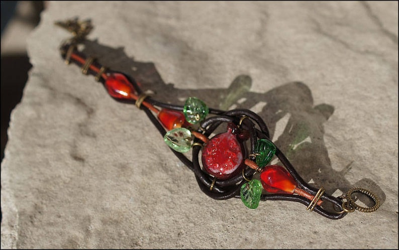 Op de afbeelding: Een leren armband met rode en groene glazen kralen, een rode glazen granaatappel en een messing sluiting.