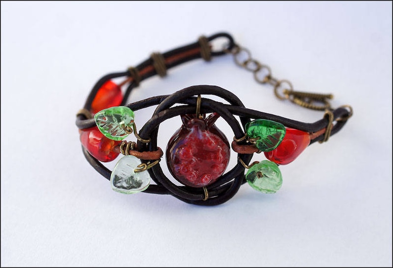 Op de afbeelding: Een bruin leren armband met een rode glazen granaatappelbedel en groene glazen bladeren.