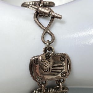 Puede incluir: Una pulsera de metal plateado con eslabones rectangulares que presentan motivos de manos en relieve y detalles en espiral. La pulsera incluye un peque&ntilde;o dije en forma de coraz&oacute;n con el n&uacute;mero 93 grabado y un cierre de barra.