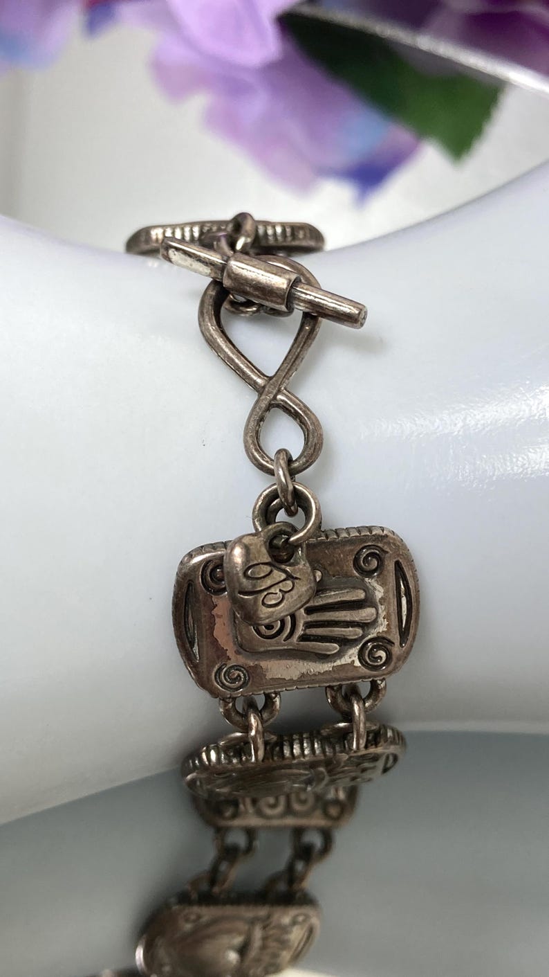 Puede incluir: Una pulsera de metal plateado con eslabones rectangulares que presentan motivos de manos en relieve y detalles en espiral. La pulsera incluye un peque&ntilde;o dije en forma de coraz&oacute;n con el n&uacute;mero 93 grabado y un cierre de barra.