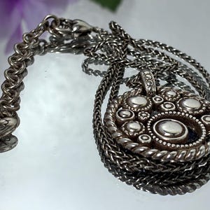 K&ouml;nnte beinhalten: Eine silberfarbene Halskette mit einem runden Anh&auml;nger, der ein strukturiertes, mehrschichtiges Design aus erhabenen Punkten und glatten Metallkreisen aufweist. Der Anh&auml;nger ist an einer geflochtenen Kette mit einem herzf&ouml;rmigen Anh&auml;nger befestigt.