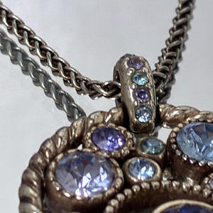 K&ouml;nnte beinhalten: Eine silberfarbene Metall-Anh&auml;nger-Halskette mit kreisf&ouml;rmigem Design und gedrehter Kordelbord&uuml;re. Der Anh&auml;nger ist mit mehreren runden, facettierten Kristallen in verschiedenen Blau-, Immergr&uuml;n- und Violettt&ouml;nen besetzt, auch an der &Ouml;se.