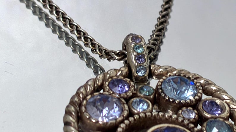 K&ouml;nnte beinhalten: Eine silberfarbene Metall-Anh&auml;nger-Halskette mit kreisf&ouml;rmigem Design und gedrehter Kordelbord&uuml;re. Der Anh&auml;nger ist mit mehreren runden, facettierten Kristallen in verschiedenen Blau-, Immergr&uuml;n- und Violettt&ouml;nen besetzt, auch an der &Ouml;se.
