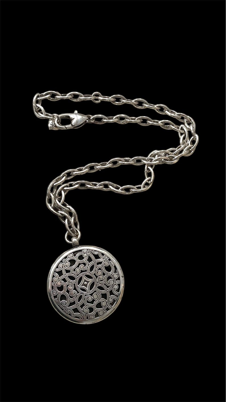 May include: Silver-toned necklace with a large, round pendant featuring an intricate openwork design. The pendant hangs from a chunky chain with a lobster clasp closure. The necklace is displayed against a black background.