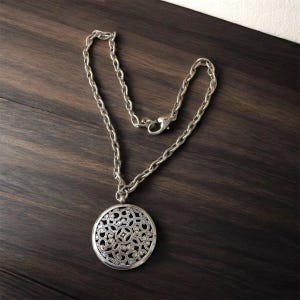 May include: Silver-toned necklace with a round pendant featuring an openwork floral design. The chain is a chunky link style with a lobster clasp closure. The pendant is approximately 2 inches in diameter.