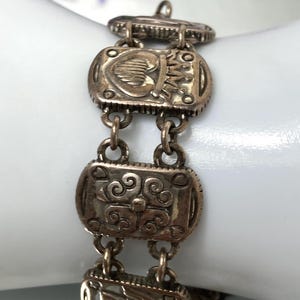 Puede incluir: Una pulsera de eslabones de metal plateado con segmentos rectangulares unidos por peque&ntilde;as anillas circulares. Cada eslab&oacute;n rectangular muestra un dise&ntilde;o en relieve distinto, incluyendo un coraz&oacute;n con corona, volutas florales y un motivo de escudo.