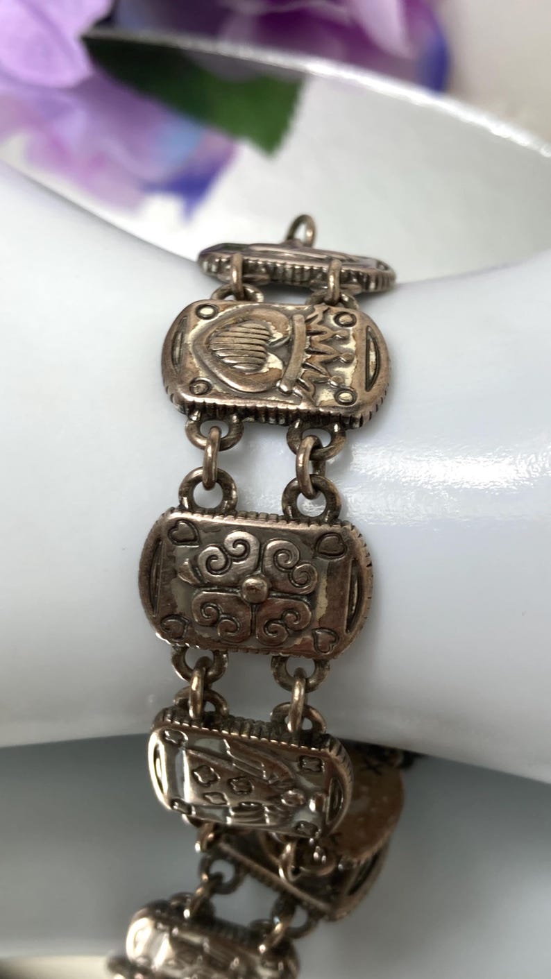 Puede incluir: Una pulsera de eslabones de metal plateado con segmentos rectangulares unidos por peque&ntilde;as anillas circulares. Cada eslab&oacute;n rectangular muestra un dise&ntilde;o en relieve distinto, incluyendo un coraz&oacute;n con corona, volutas florales y un motivo de escudo.