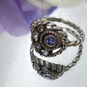 May include: A silver-toned ring with a central purple gemstone and smaller clear and blue stones. The ring has a detailed, textured design with a twisted band. The ring is reflected on a shiny surface, with a blurred purple flower in the background.