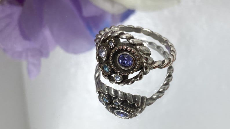 May include: A silver-toned ring with a central purple gemstone and smaller clear and blue stones. The ring has a detailed, textured design with a twisted band. The ring is reflected on a shiny surface, with a blurred purple flower in the background.