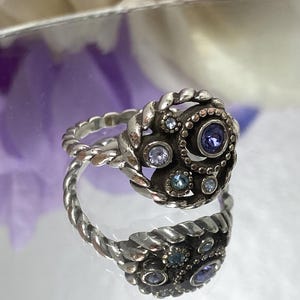 May include: Silver-toned ring with a twisted band and a cluster of small, round, and oval gemstones in shades of blue and purple. The ring's centerpiece features a larger, dark purple gemstone. The ring is displayed on a reflective surface.