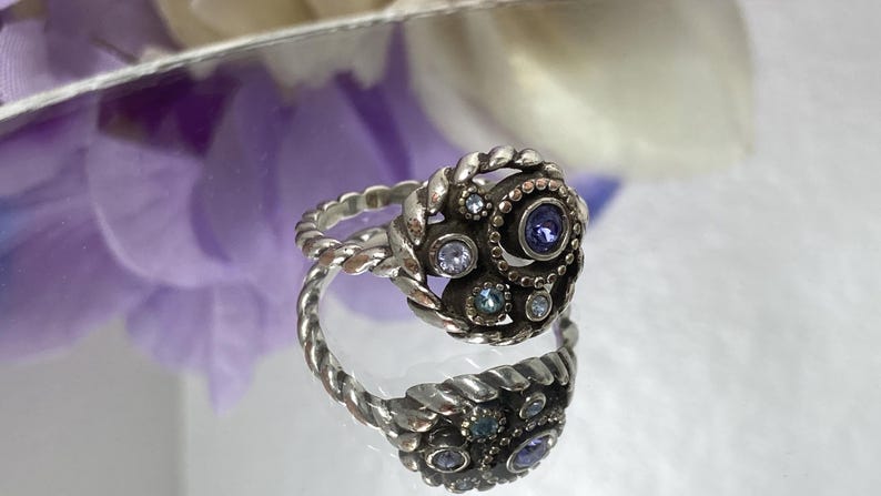May include: Silver-toned ring with a twisted band and a cluster of small, round, and oval gemstones in shades of blue and purple. The ring's centerpiece features a larger, dark purple gemstone. The ring is displayed on a reflective surface.
