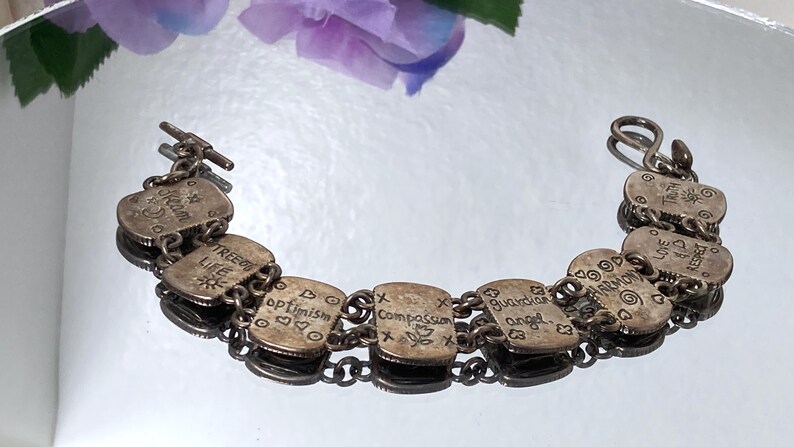 Puede incluir: Una pulsera de eslabones de metal plateado con siete dijes rectangulares. Cada dije est&aacute; grabado con palabras como Dream, Tree of Life, Optimism, Compassion, Guardian Angel, Harmony, Love and Respect y Truth, junto a peque&ntilde;os s&iacute;mbolos.