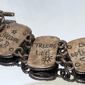 Puede incluir: Una pulsera de eslabones de metal plateado con dijes rectangulares grabados con texto y s&iacute;mbolos. Los dijes muestran las palabras dream, TREE OF LIFE y optimism, acompa&ntilde;adas de estrellas, una luna creciente, un sol y formas de coraz&oacute;n.