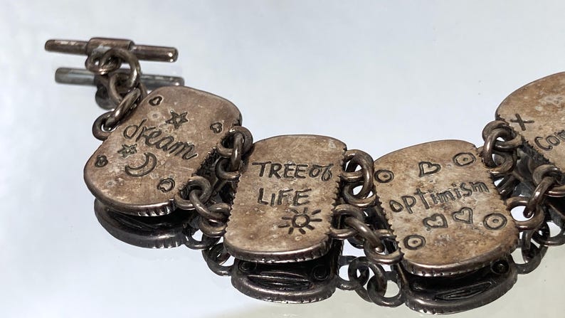 Puede incluir: Una pulsera de eslabones de metal plateado con dijes rectangulares grabados con texto y s&iacute;mbolos. Los dijes muestran las palabras dream, TREE OF LIFE y optimism, acompa&ntilde;adas de estrellas, una luna creciente, un sol y formas de coraz&oacute;n.