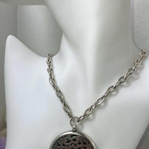 May include: Silver-toned necklace with a large, round pendant. The pendant features an intricate openwork design over a dark background. The chain is a chunky link style, displayed on a white mannequin bust. A stylish accessory.