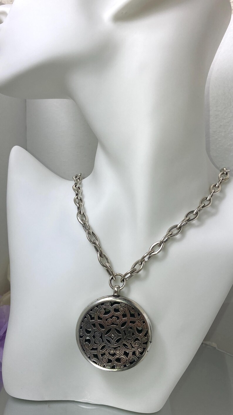 May include: Silver-toned necklace with a large, round pendant. The pendant features an intricate openwork design over a dark background. The chain is a chunky link style, displayed on a white mannequin bust. A stylish accessory.