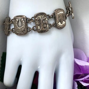 Puede incluir: Una pulsera de eslabones de metal plateado con dijes rectangulares que presentan dise&ntilde;os en relieve, incluyendo una herradura, un &aacute;rbol y un coraz&oacute;n. La pulsera se asegura con un cierre de barra y consta de varios segmentos met&aacute;licos unidos.