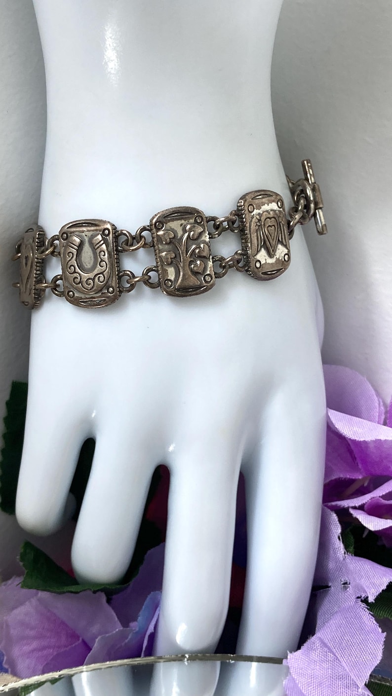 Puede incluir: Una pulsera de eslabones de metal plateado con dijes rectangulares que presentan dise&ntilde;os en relieve, incluyendo una herradura, un &aacute;rbol y un coraz&oacute;n. La pulsera se asegura con un cierre de barra y consta de varios segmentos met&aacute;licos unidos.