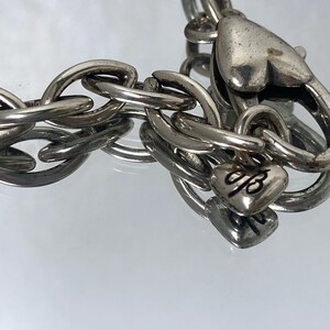 May include: Silver-toned chain bracelet with large oval links. The bracelet features a heart-shaped clasp and a small heart charm with an engraved design. The jewelry is displayed on a reflective surface.