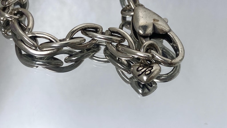 May include: Silver-toned chain bracelet with large oval links. The bracelet features a heart-shaped clasp and a small heart charm with an engraved design. The jewelry is displayed on a reflective surface.