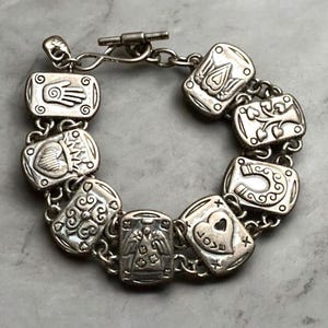 Puede incluir: Una pulsera de dijes en tono plateado con ocho eslabones rectangulares, cada uno grabado con s&iacute;mbolos distintos que incluyen una mano, un coraz&oacute;n, un &aacute;rbol, una herradura y un &aacute;ngel. La pulsera se asegura con un cierre de barra y eslabones circulares.