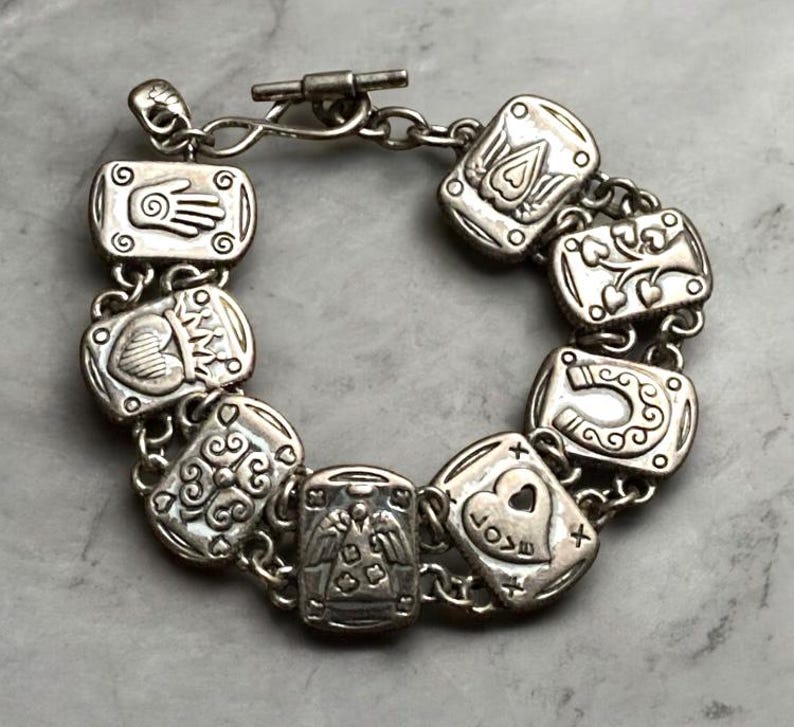 Puede incluir: Una pulsera de dijes en tono plateado con ocho eslabones rectangulares, cada uno grabado con s&iacute;mbolos distintos que incluyen una mano, un coraz&oacute;n, un &aacute;rbol, una herradura y un &aacute;ngel. La pulsera se asegura con un cierre de barra y eslabones circulares.