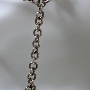 K&ouml;nnte beinhalten: Eine silberfarbene Halskette mit geflochtenem Kettenmuster, Karabinerverschluss und einer Verl&auml;ngerungskette aus runden Gliedern. Ein kleiner, herzf&ouml;rmiger Anh&auml;nger mit gepr&auml;gtem Design h&auml;ngt am Ende der Verl&auml;ngerungskette.