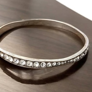 Puede incluir: Una pulsera r&iacute;gida de metal en tono plateado que presenta una hilera continua de cristales transparentes de corte redondo incrustados en la superficie exterior. La pulsera tiene un acabado liso y pulido con una forma circular.